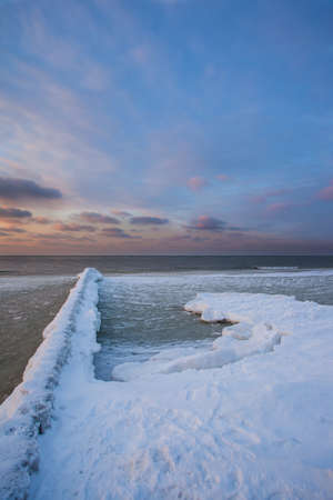 snow-covered breakwaters on a winter beach near a cold, ice-covered sea during sunsetの写真素材