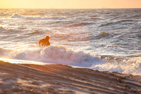 silhouette of a man in a diving suit with a net on the beach collecting amber in the water against the backdrop of a bright orange sunsetの写真素材
