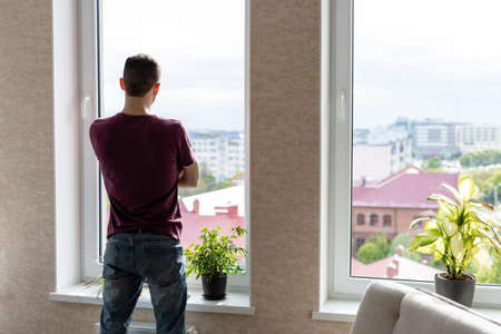 young man in a t-shirt and jeans stands at a large window and looks at the city