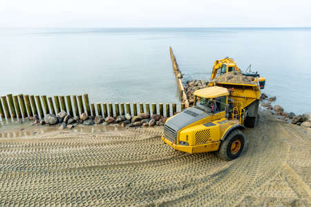an excavator on the beach loads sand and stones into the truck body, works to strengthen the beach and install breakwatersの写真素材