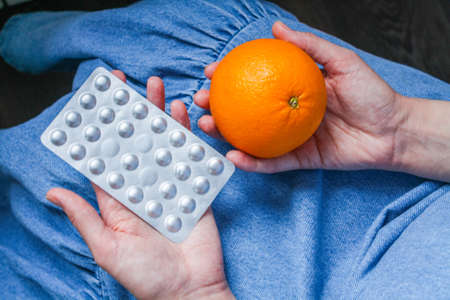 A young woman holds her hands in her lap, palms up. On the open palms are an orange, a pear, blisters, tablets, pillsの写真素材