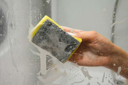 A womans hand with a yellow sponge washes the glass in the shower stallの写真素材
