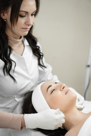 Clinical purity: in the beauty clinic, a cosmetologist cleanses a womans face, providing her with a healthy and radiant look. The patient is a young woman at the reception at the cosmetologist.の写真素材