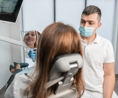 A female patient looks in a mirror showing her a dentist in a clinic. A woman in the clinic receives a full range of dental services, from treatment to teeth alignment.の写真素材