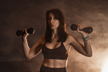 A Female Fitness Model is actively engaging with dumbbells in a welllit studio setting, showcasing her dedication to maintaining a healthy lifestyle through vigorous and effective exercise routinesの写真素材