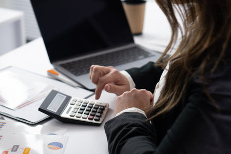 A dedicated business professional is diligently calculating financial data utilizing a calculator, while surrounded by paperwork, a laptop, a coffee mug, and an atmosphere conducive to concentrationの写真素材