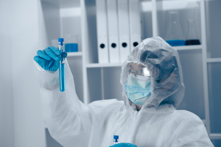 A scientist clad in protective gear is meticulously analyzing a liquid sample contained within a laboratory test tube, demonstrating the critical role of safety and precision in scientific researchの写真素材