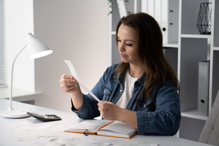 A focused woman meticulously analyzes her receipts at an organized desk, showcasing her attention to detail and highlighting the importance of careful financial management and organization in lifeの写真素材