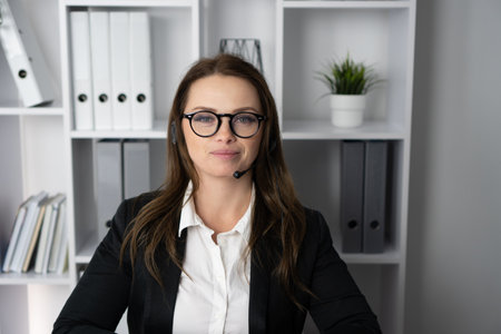 A confident professional woman in a headset is in a modern office, focusing on business communication. Surrounded by folders, a laptop, and a plant, she represents success and teamwork with a smileの写真素材