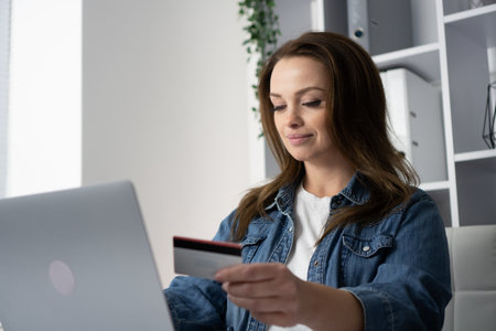 A woman sits at her desk, engaged in online shopping on her laptop, browsing products and making transactions with her credit card, highlighting the ease of modern consumer habitsの写真素材