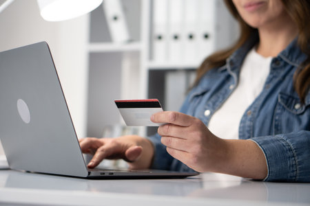 A young woman engages in online shopping, actively using her credit card while sitting at her desk with a laptop. This modern consumer experience exemplifies convenience and the digital lifestyleの写真素材