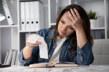 A Frustrated Woman is Thoroughly Reviewing Receipts and Financial Records at Her Desk, Trying to Manage Her Bills and Business Expenses While Feeling Overwhelmed and Concerned About Her Financesの写真素材