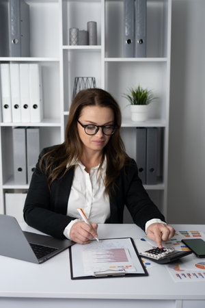 In a modern office setting, a businesswoman is meticulously analyzing financial data while using her laptop and consulting various charts and reports on her desktop to ensure productivity and successの写真素材