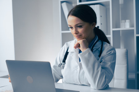 A dedicated and focused female doctor is thoroughly reviewing important patient information using her laptop, demonstrating her commitment to providing optimal healthcare and thoughtful patient careの写真素材