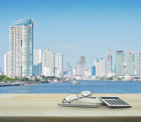 Pen, glasses, notebook and calculator on wooden table with city, business backgroundの写真素材