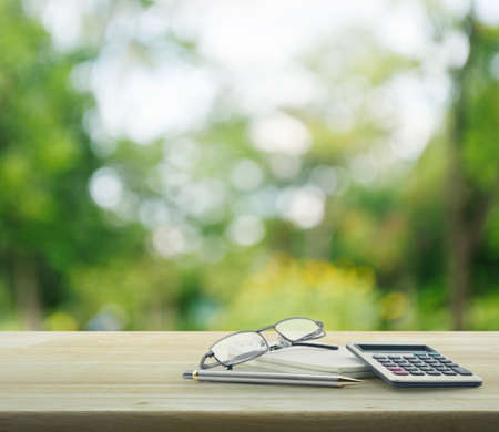 Pen, glasses, notebook and calculator on wooden table over green tree bokeh backgroundの写真素材