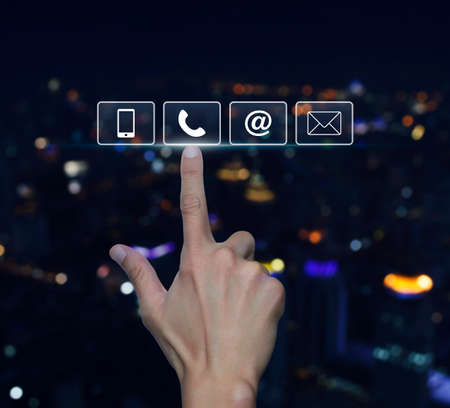 Hand pressing telephone, mobile phone, at and email buttons over blurred light city tower...