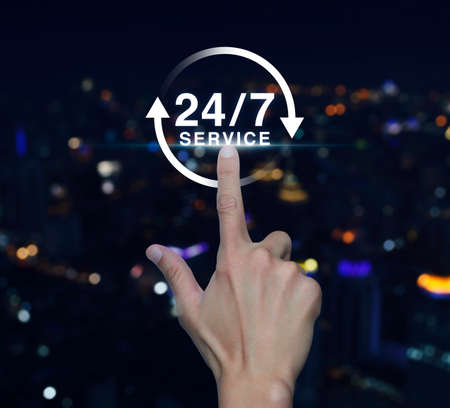 Hand pressing button 24 hours service icon over blurred light city tower background, Full time service conceptの写真素材
