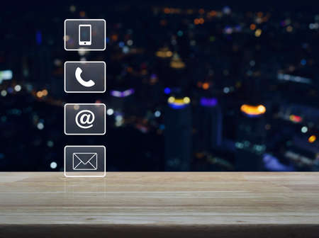 Telephone, mobile phone, at and email buttons on wooden table in front of blurred light city tower, Customer support conceptの写真素材