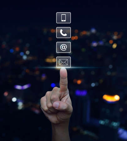 Hand pressing telephone, mobile phone, at and email buttons over blurred light city tower background, Customer support conceptの写真素材