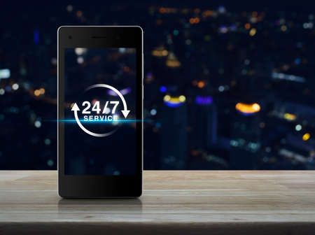 24 hours service icon on modern smart phone screen on wooden table over blur light city tower...