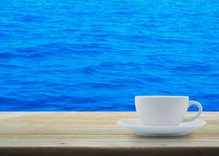 White cup on wooden table over blue sea water surface textureの写真素材