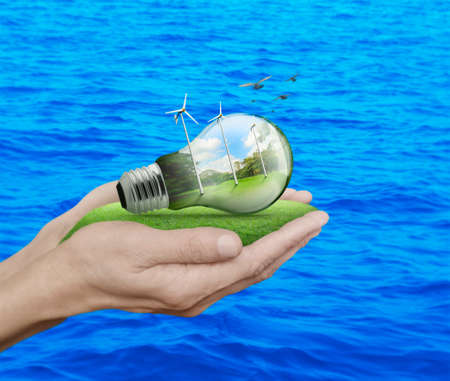 Light bulb with wind turbines, birds and forest inside in hands over blue sea background, Ecological conceptの写真素材