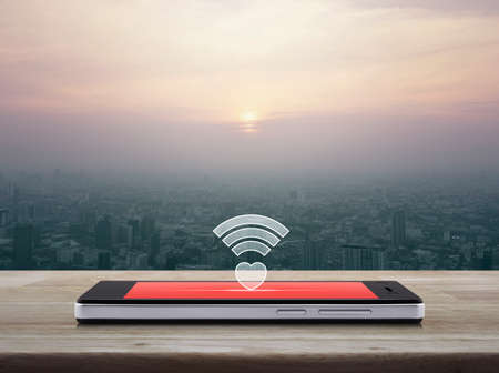 Heart wifi icon on modern smart phone screen on wooden table over aerial view of cityscape at...