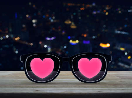 Love heart with eye glasses on wooden table over blur colourful night light city tower, Valentines day conceptの写真素材