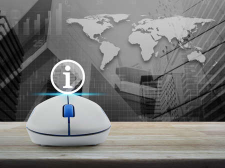Wireless computer mouse with information sign icon on wooden table over world map with financial graph and city towerの写真素材