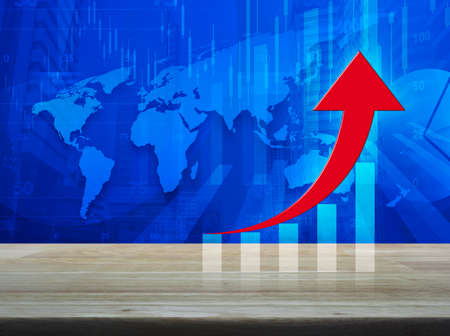 Red arrow with graph on wooden table over world map with financial graph and city tower background,...