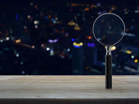 Magnifying glass on wooden table over blur colorful night light city tower background, Business analyzing conceptの写真素材