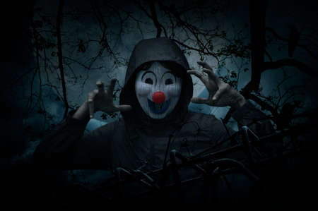 Scary evil clown wear jacket with old fence over dead tree, crow, moon and spooky cloudy sky, Halloween conceptの写真素材