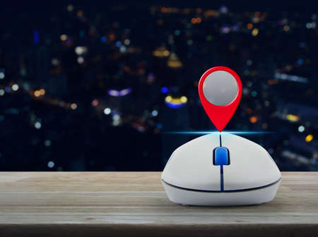 Map pin location button with wireless computer mouse on wooden table over blur colorful night light city tower, Map pointer navigation conceptの写真素材
