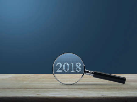 Magnifying glass with 2018 text and graph on wooden table over light blue gradient background,...