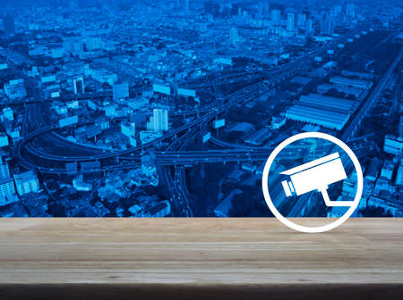 cctv camera icon on wooden table over modern city tower, street and expressway, Business security...