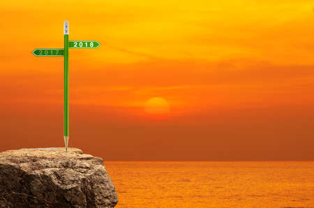 2018 and 2017 direction sign plate with green pencil on rock over sunset sky and sea, Business success strategy plan concept, Happy new year 2018 calendar coverの写真素材