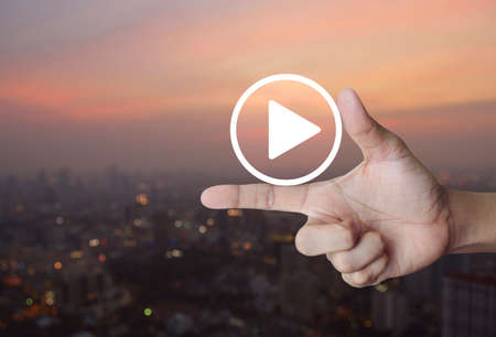 Play button icon on finger over blur of cityscape on warm light sundown, Business music online...