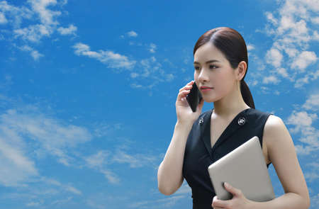 Young beautiful asian businesswoman talking on smart mobile phone over blue sky with white clouds, Business communication conceptの写真素材