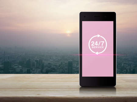24 hours service icon on modern smart phone screen on wooden table over city tower at sunset,...
