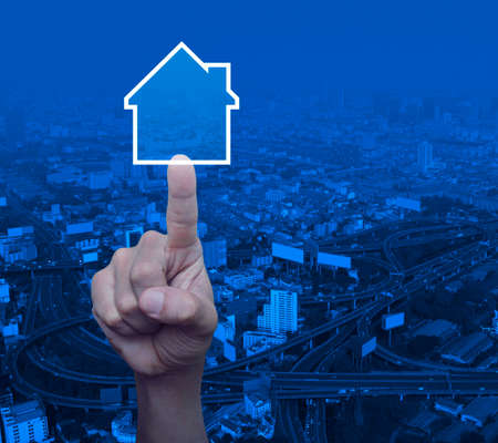 Hand pressing house icon with copy space over modern city tower, street and expressway, Real estate...
