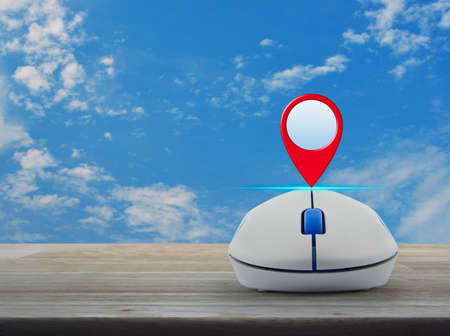 Map pin location button with wireless computer mouse on wooden table over blue sky with white...