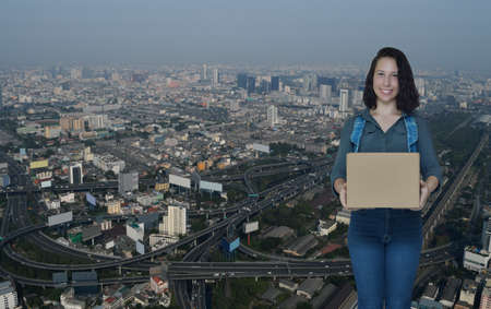 Smiling young caucasian woman holding parcel box over modern city tower, street and expressway and...