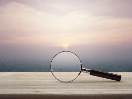 Magnifying glass on wooden table over modern city tower building and skyscraper at sunset, vintage style, Business analyzing conceptの写真素材