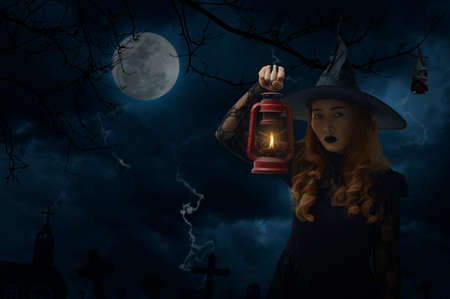 Halloween witch holding ancient lamp standing over dead tree, bat, full moon and cloudy sky, Halloween mystery conceptの写真素材