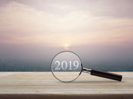 Magnifying glass with 2019 text and graph on wooden table over modern city tower building and skyscraper at sunset, vintage style, Business happy new year research conceptの写真素材