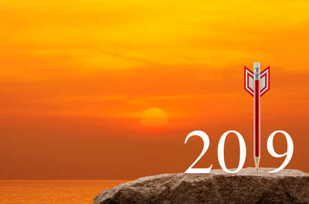 Red pencil in the shape of a dart arrow with 2019 white text on rock mountain over sunset sky and...