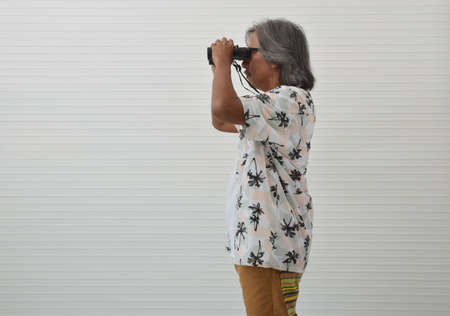 Senior traveler asian man wearing glasses, summer shirt and brown shorts holding binoculars over...