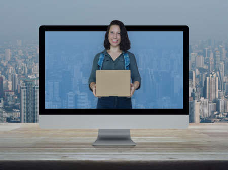 Smiling young caucasian woman holding parcel box on desktop modern computer monitor screen on wooden...