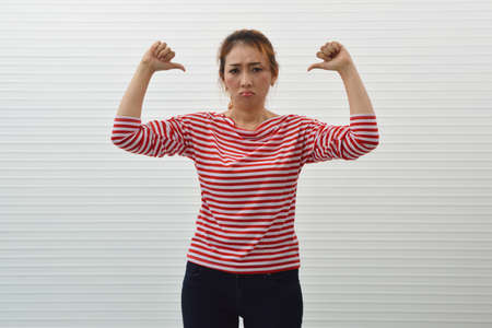 Moody young asian woman wearing red and white stripped shirt with jeans showing dislike thumps down hand over white wall background, Upset facial expressionの写真素材